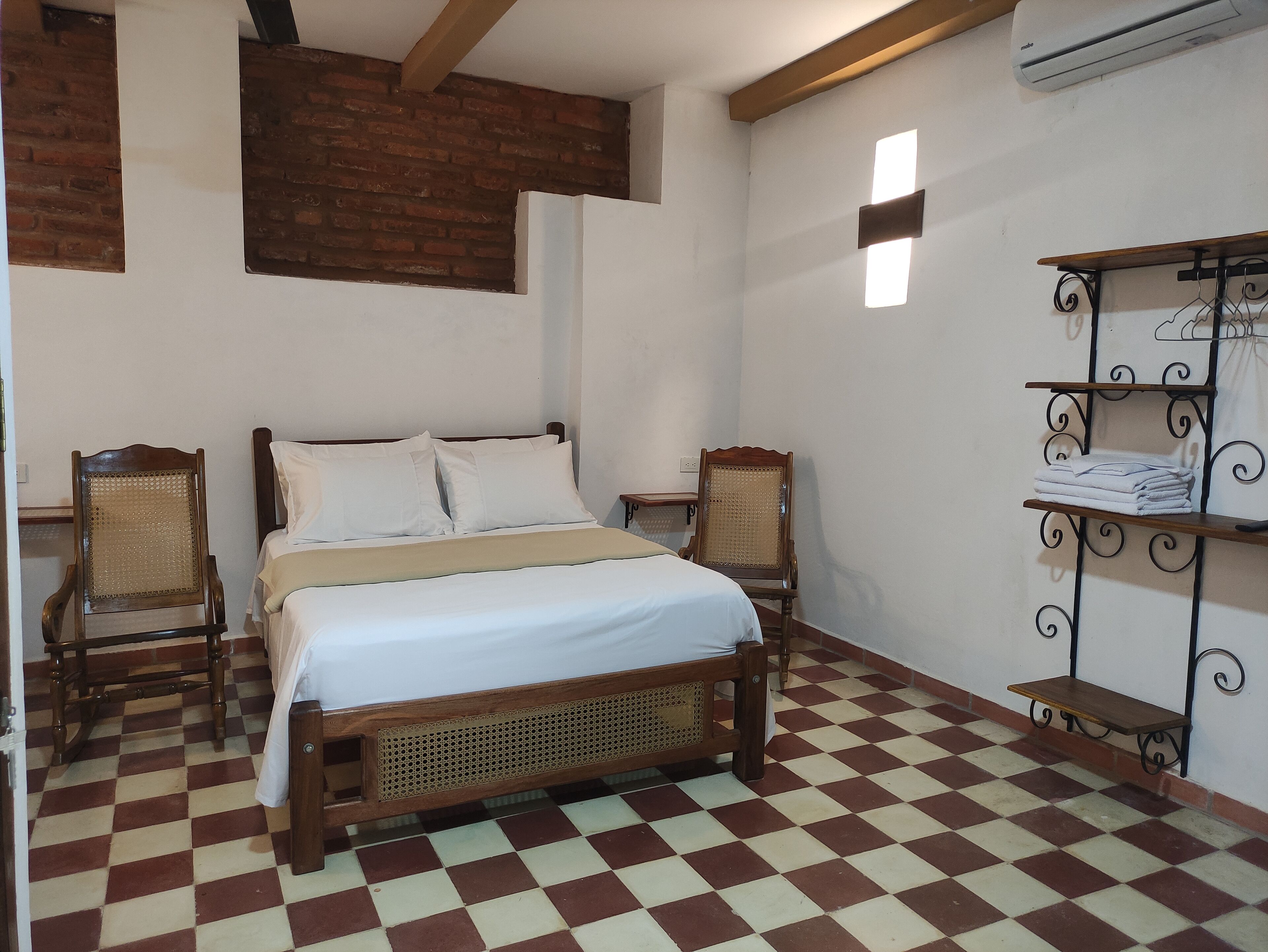 A room with a checkered floor, a wooden bed with white bedding, two wooden chairs, a metal shelf with folded towels, and a brick wall.