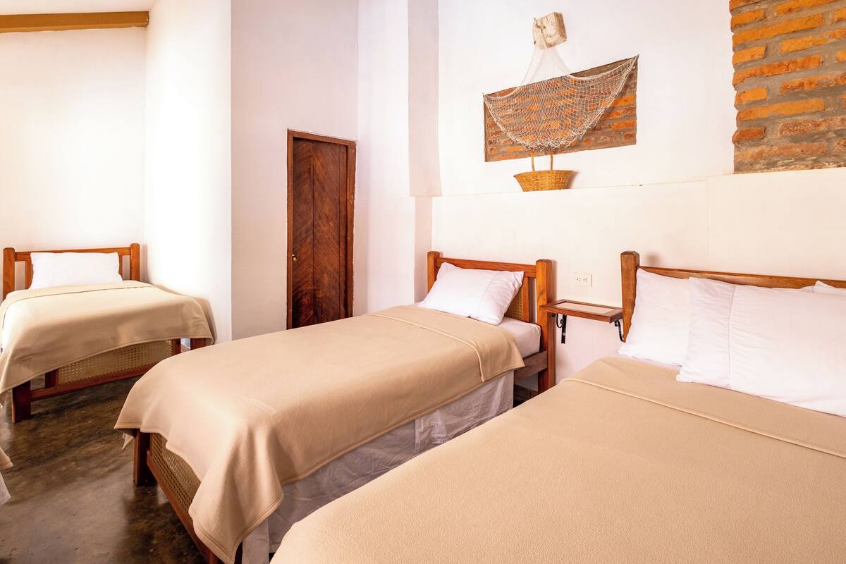 A room with three wooden beds, each with a beige bedspread and white pillows. There is a brick wall and a wooden door.
