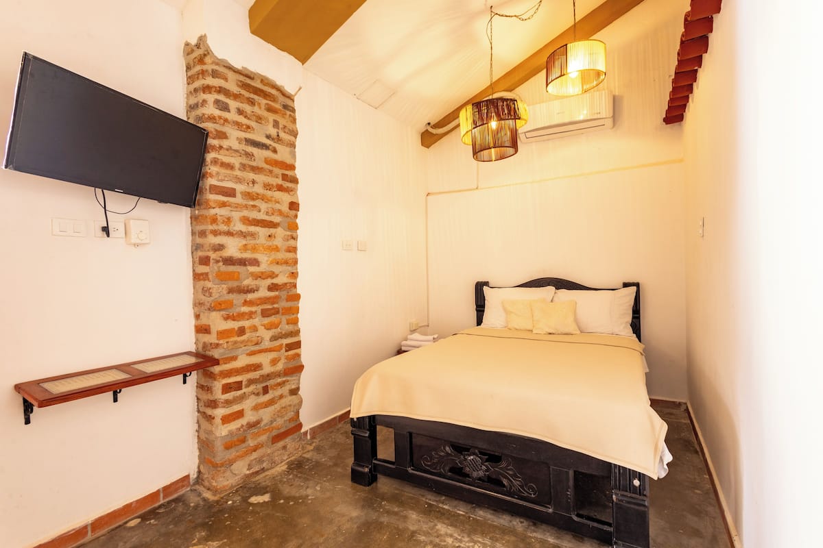 A bedroom with a brick wall, a bed with a white bedspread, a mounted TV, and a hanging light fixture.