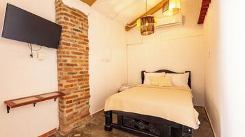 Un dormitorio con pared de ladrillo, cama con cubrecama blanco, televisor montado y lámpara colgante.