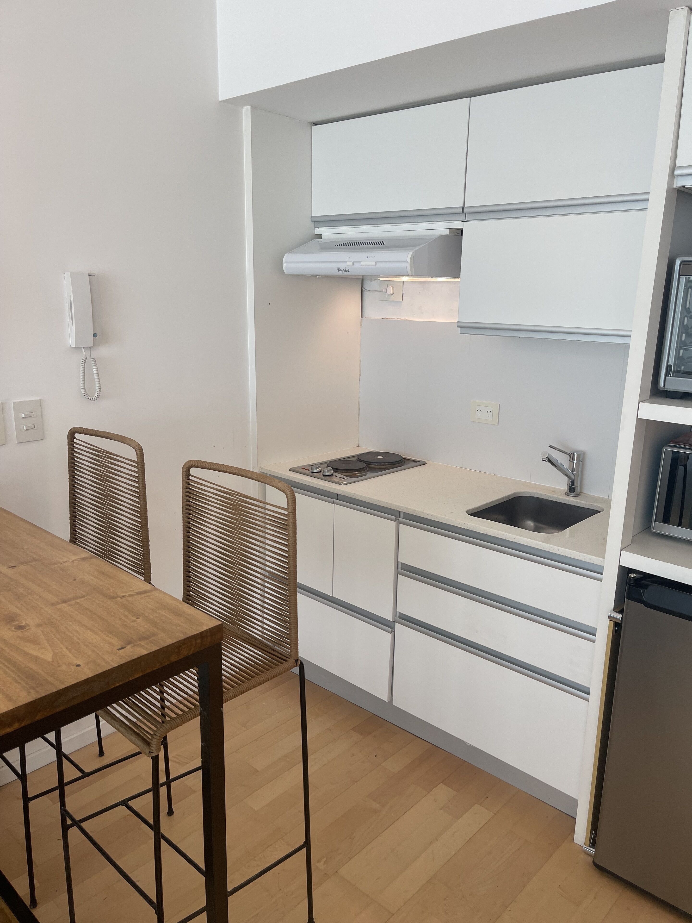 Una cocina compacta con alacenas blancas, horno integrado y un lavamanos. Hay una mesa de comedor de madera y dos sillas de mimbre.