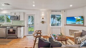 Uma cozinha moderna com armários brancos e eletrodomésticos de aço inoxidável, uma sala de jantar com mesa e cadeiras de madeira, e uma sala de estar com sofá e televisão exibindo uma cena de praia.