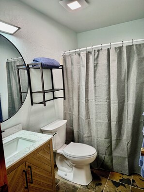 Un bany amb vàter, un lavabo amb taulell de marbre, mirall, penjador de tovalloles i cortina de bany.
