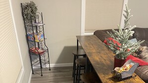 Uma área de refeições com uma mesa e cadeiras de madeira, uma estante de metal com caixas, uma planta em vaso e uma pequena árvore decorativa.