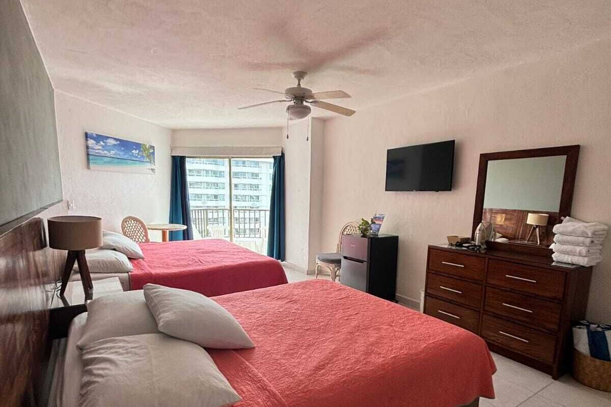 A hotel room with two beds, a ceiling fan, a TV, a dresser, and a window with a view of buildings.
