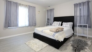 Une chambre avec une tête de lit noire, un lit avec une literie blanche, des rideaux gris, un tapis blanc et un tapis à motifs zébrés.