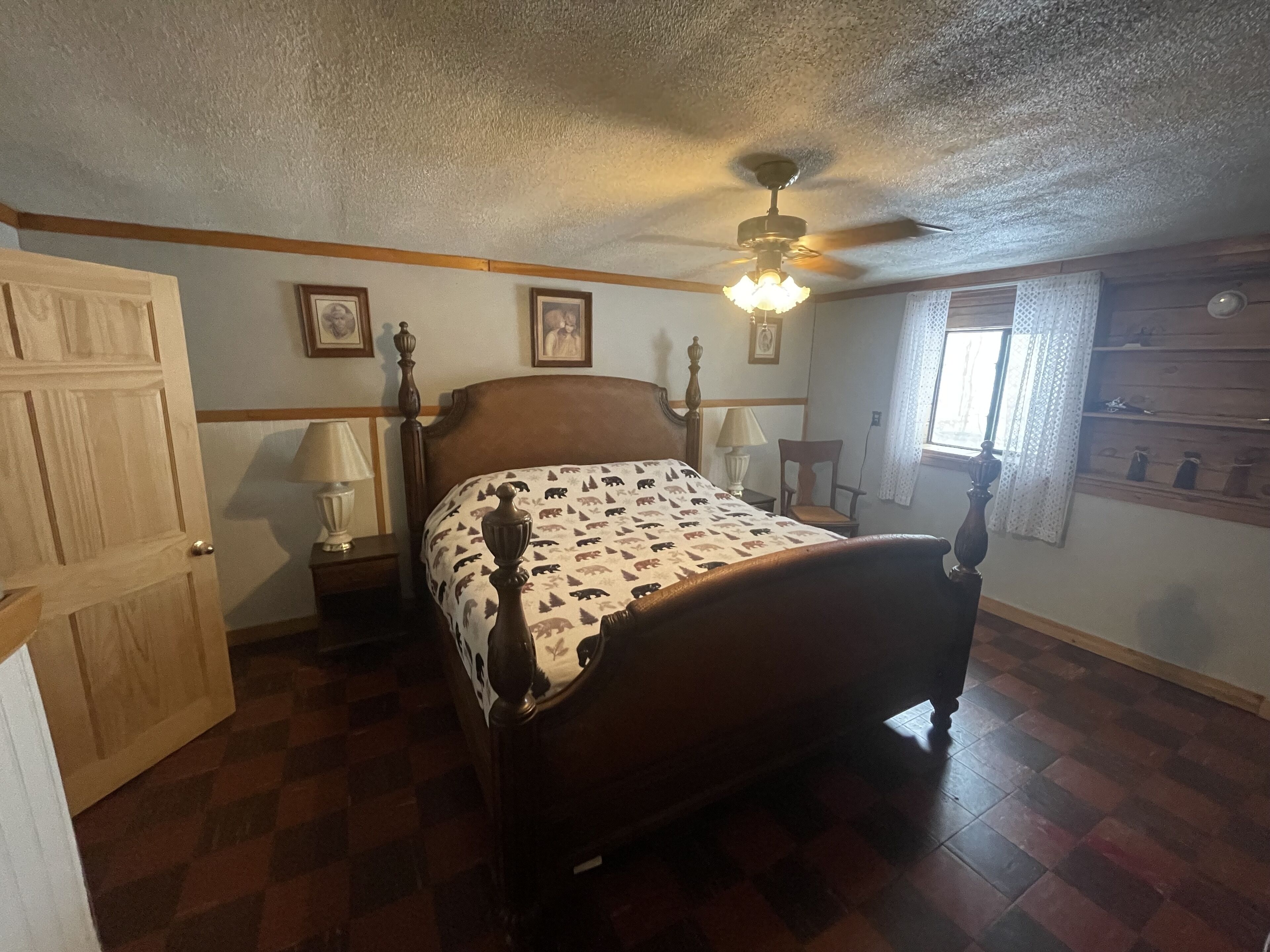 Un dormitorio con una cama de madera, un ventilador de techo, una ventana con cortinas de encaje y un aparador de madera.
