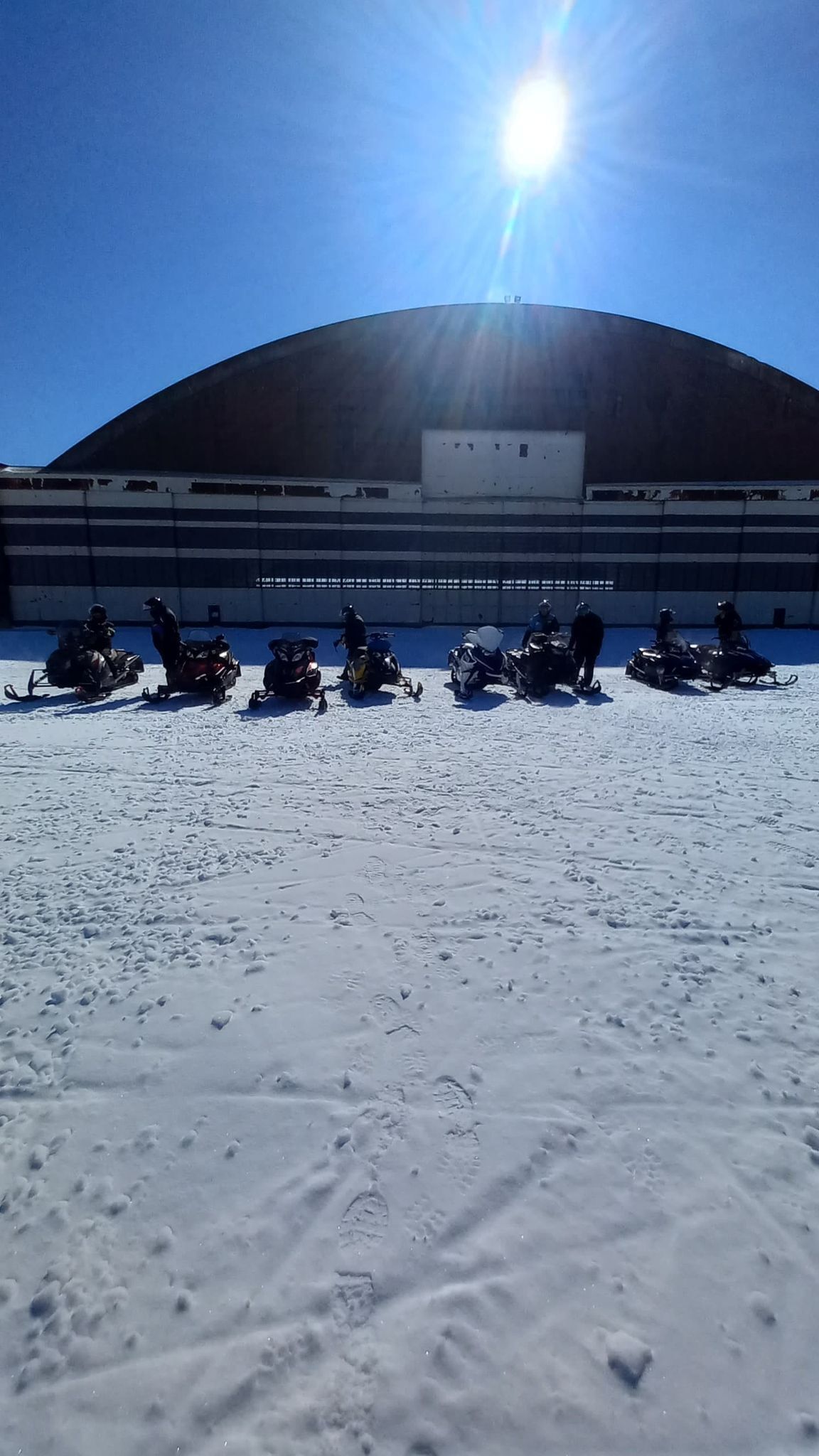 A large indoor space with a curved roof, possibly a hangar or warehouse, with several snowmobiles parked in the foreground.