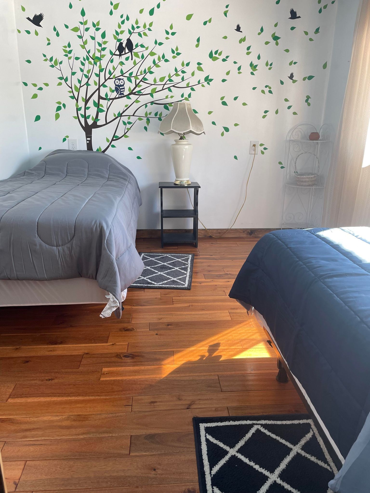 A bedroom with two beds, a nightstand with a lamp, a wall-mounted shelf, and a decorative tree decal.