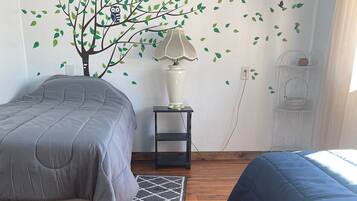 A bedroom with two beds, a nightstand with a lamp, a wall-mounted shelf, and a decorative tree decal.