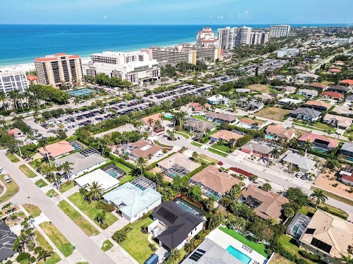 An aerial view of a coastal residential area with a mix of single-family homes, a hotel, and a beach in the background.