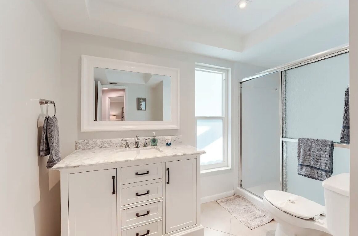 A modern bathroom with a white vanity, a large mirror, a shower enclosure, and a toilet.