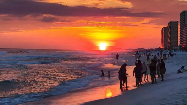 Une scène de plage où l’on voit des personnes se promener et se tenir debout sur le sable, avec des bâtiments en arrière-plan.