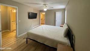 Une chambre à coucher comprenant un lit, un ventilateur de plafond, un téléviseur fixé au mur et une porte donnant sur une autre pièce.