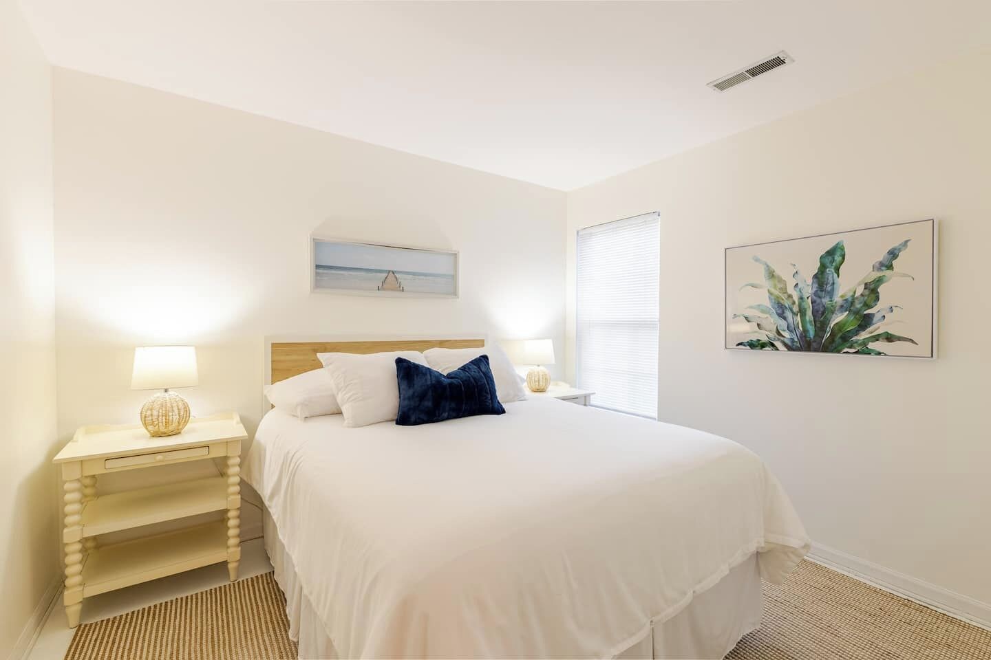 Una camera da letto con un letto, due comodini con lampade, una finestra con tende, un quadro appeso al muro e una bocchetta di ventilazione a soffitto.