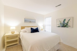 A bedroom with a bed, two bedside tables with lamps, a window with blinds, a wall-mounted picture, and a ceiling vent.