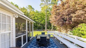 A covered outdoor deck with a table and chairs, a white railing, and a view of a backyard with trees and a hammock.