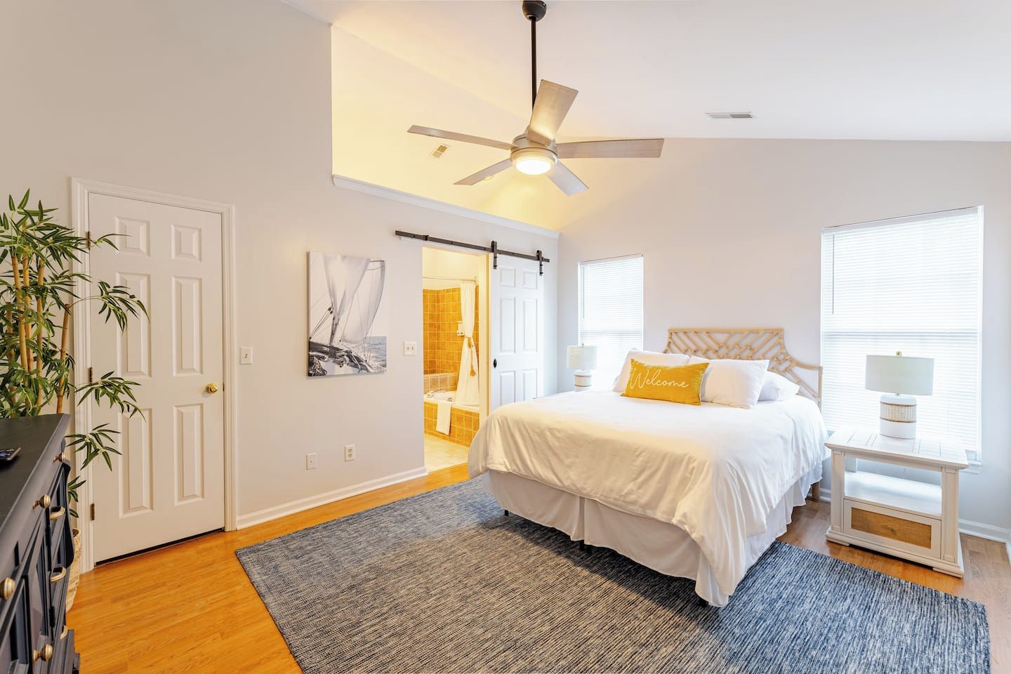 A bedroom with a bed, a ceiling fan, a nightstand, a lamp, a window, and a door leading to a bathroom.