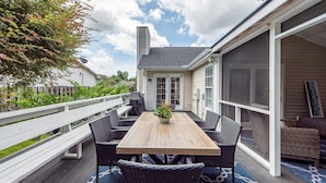 An outdoor patio with a wooden table and wicker chairs, a white fence, and a view of a house in the background.