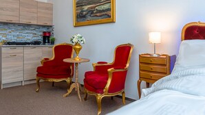 Ett hotellrum med en säng, två röda sammetsstolar, ett litet bord med en vas, en tavla på väggen, ett nattduksbord med en lampa och ett litet pentry med en kaffemaskin.