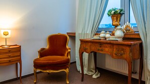 Una habitación con un sillón tapizado en terciopelo rojo, un escritorio de madera con un florero y una lámpara en una mesita de noche.