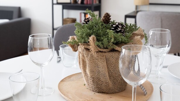 Una mesa de comedor dispuesta con copas de vino, platos y un centro de mesa con piñas y follaje dentro de un saco de arpillera.