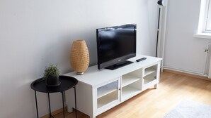 Una sala de estar moderna con un televisor de pantalla plana sobre un mueble blanco, una mesita auxiliar negra con una planta en maceta y una lámpara de pie alta.