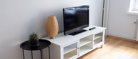 Uma sala de estar moderna com uma TV de tela plana sobre um móvel branco, uma pequena mesa lateral preta com uma planta em vaso e um abajur de chão alto.