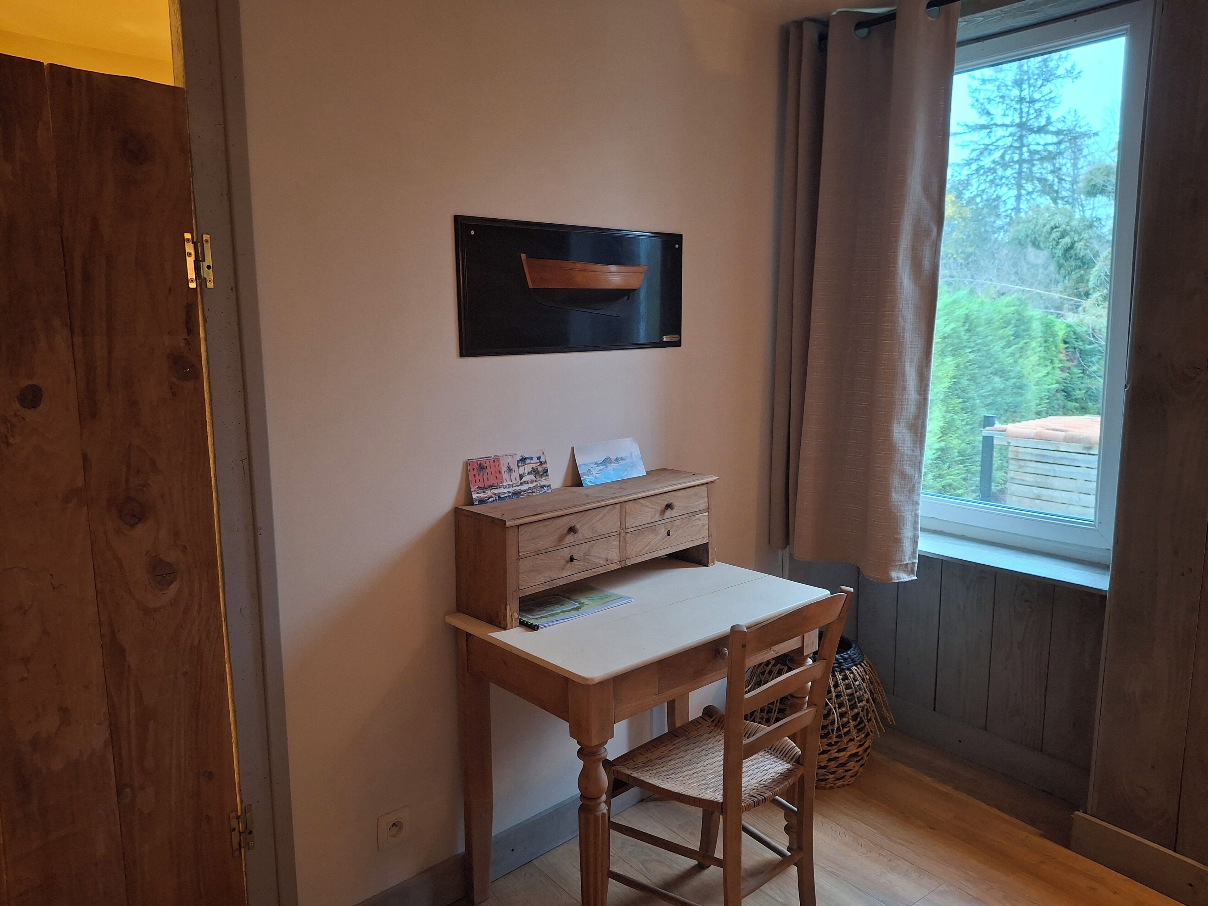 A small study room with a wooden desk, a chair, a wall-mounted TV, and a window with curtains.