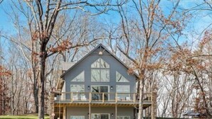 Une grande maison à deux étages, dotée d’un balcon imposant, entourée d’arbres et d’une cour verdoyante.