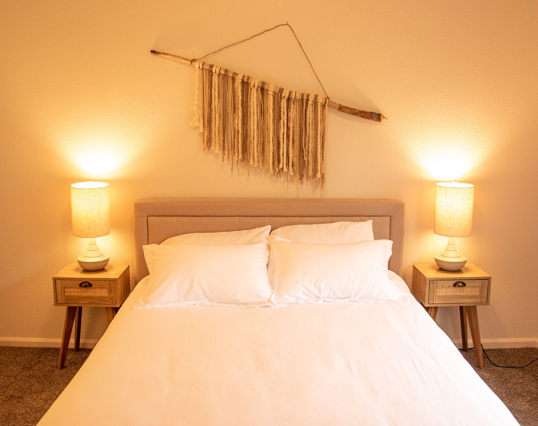 Cama com roupa de cama branca, duas luminárias de cabeceira e um painel de macramé fixado na parede.