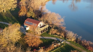 Una casa grande y antigua con techo rojo y un balcón, rodeada de árboles y un lago.