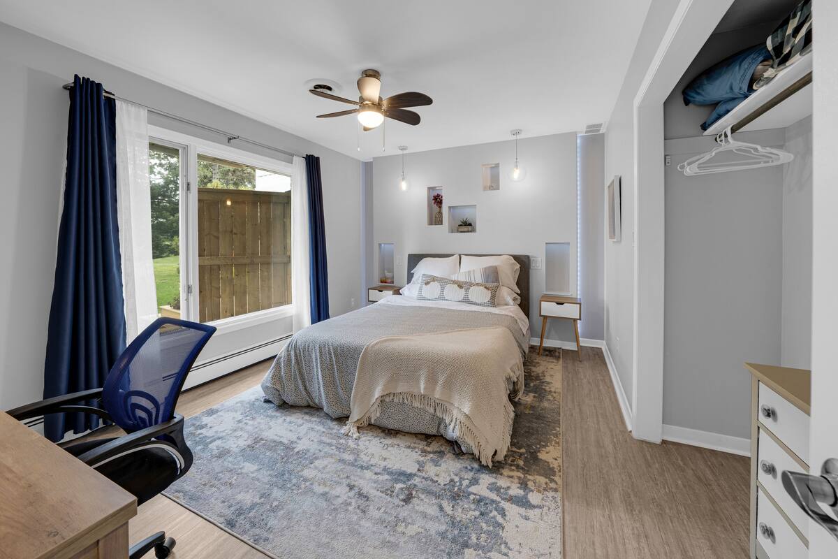 A bedroom with a bed, a desk, a chair, a ceiling fan, and a view of the outdoors.
