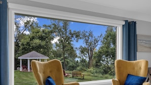 Una habitación con dos sillones color mostaza con cojines azules, una mesita blanca y un ventanal que ofrece vistas a un jardín con un pabellón.
