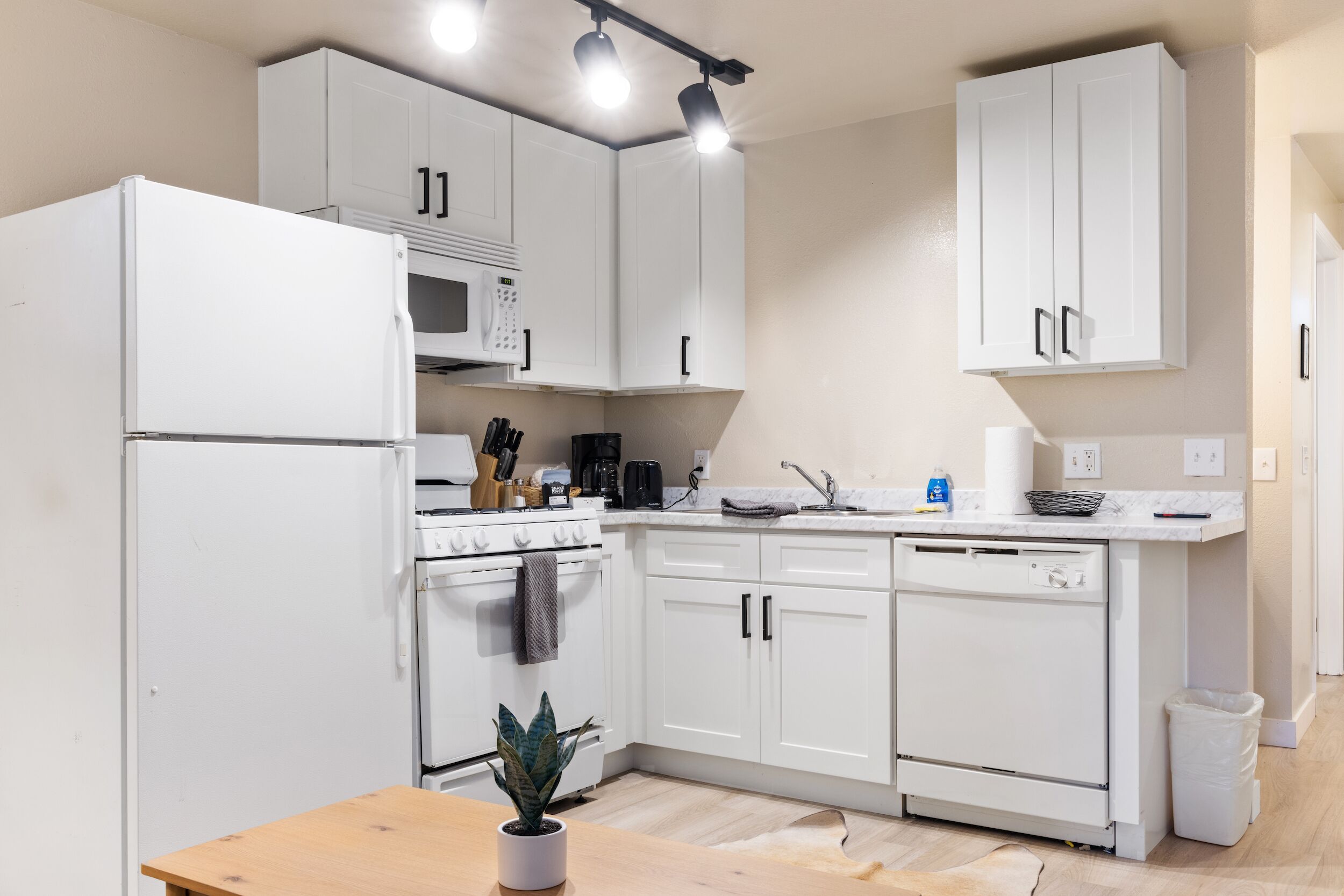 Cocina moderna con muebles blancos, microondas, estufa y lavavajillas. Hay una mesa de madera con una planta y un cubo de basura.