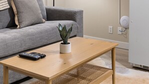 Una sala de estar con un sofá gris, una mesa de centro de madera, una planta en maceta, un televisor de pantalla plana montado en la pared y un refrigerador blanco.