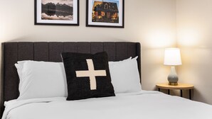 Una cama bien tendida con ropa blanca, cabecera oscura y almohada negra con una cruz blanca. Hay dos cuadros enmarcados en la pared, una lámpara sobre una mesita de madera y una pequeña mesa redonda con una lámpara.