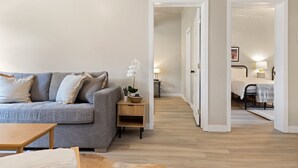 A modern living room with a grey sofa, a small wooden table, and a potted orchid. A hallway leads to a bedroom with a bed and a nightstand. - Outpost: Town Square Lodge 8 - Close to Town Square (Jackson)