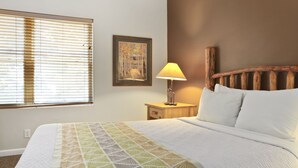 A neatly made bed with a geometric patterned bedspread, a bedside lamp, a framed artwork on the wall, and a window with blinds. - Outpost: Town Square Lodge 9 - Close to Town Square (Jackson)