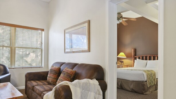 A room with a brown leather sofa, a wooden table, a bed with a white and green bedspread, and a ceiling fan. - Outpost: Town Square Lodge 9 - Close to Town Square (Jackson)