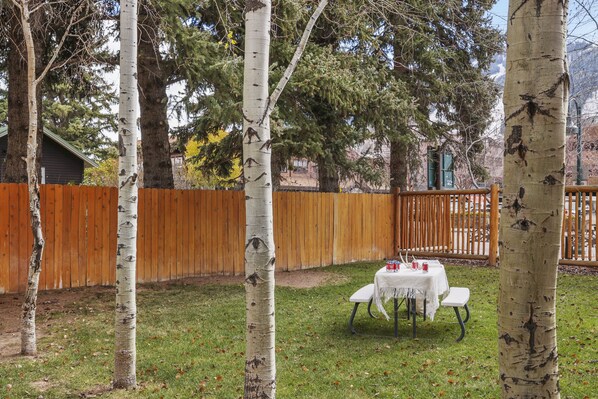 Patio con abedules, cerca de madera y mesa de picnic con mantel blanco y vasos rojos.