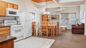 A kitchen with wooden cabinets, a white stove, and a microwave. There is a dining area with a table and chairs, and a living room with a couch and a lamp.