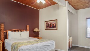 A bedroom with a wooden headboard, a bed with a quilt, a nightstand with a lamp, a framed picture on the wall, and a window with blinds.