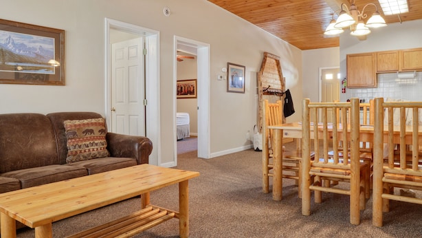 A cozy living area with a wooden table and chairs, a brown leather couch with a patterned pillow, a white door, and a kitchen area with wooden cabinets.