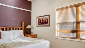 Ein Schlafzimmer mit Bett, Nachttisch, Lampe, einem gerahmten Bild an der Wand und einem Fenster mit Jalousien.