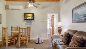 Ein Wohnzimmer mit Holzbalkendecke, Deckenventilator, Wand-Fernseher, braune Ledersofa mit gemusterten Kissen, Esstisch mit Stühlen und einer weißen Tasche auf dem Boden.