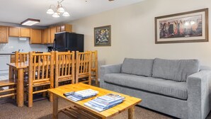 Un salon avec un canapé gris, une table à manger en bois avec des chaises, un réfrigérateur noir, des meubles en bois, un ventilateur de plafond et un tableau encadré accroché au mur.