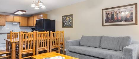 A living room with a grey sofa, wooden dining table with chairs, a black refrigerator, wooden cabinets, a ceiling fan, and a framed picture on the wall.