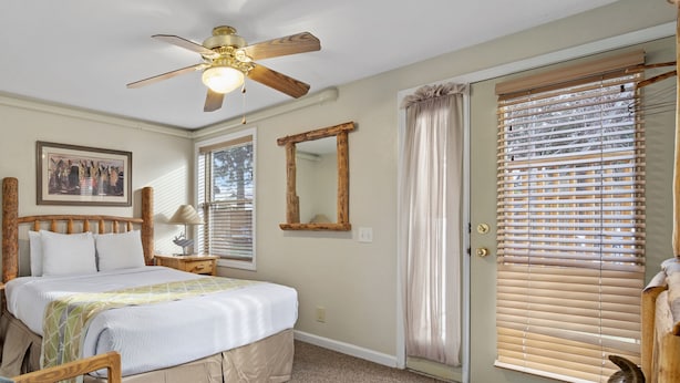 A bedroom with a bed, a nightstand, a chair, a ceiling fan, a window with blinds, and a door with a mirror. - Outpost: Town Square Lodge 6 - Close to Town (Jackson)