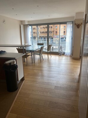A spacious room with a large dining table and chairs, a trash bin, and a view of a building with balconies through large windows.